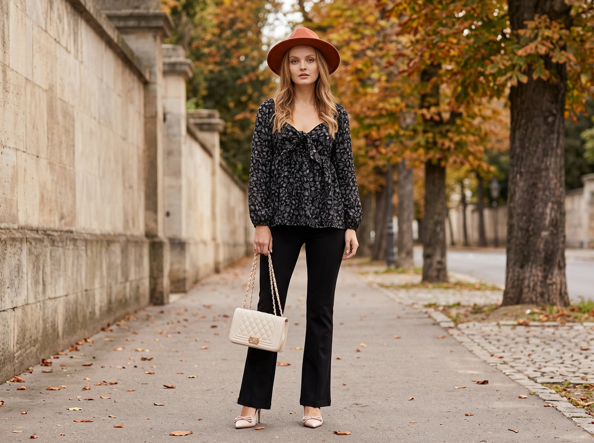A woman in a black printed tie-front babydoll blouse, black flare pants and nude pumps with a cream quilted bag