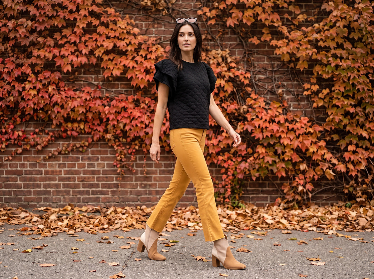 A woman in a black quilted puff-sleeve top, mustard kick flare pants and tan booties, sunglasses on her head, walks past a brick wall covered in red ivy