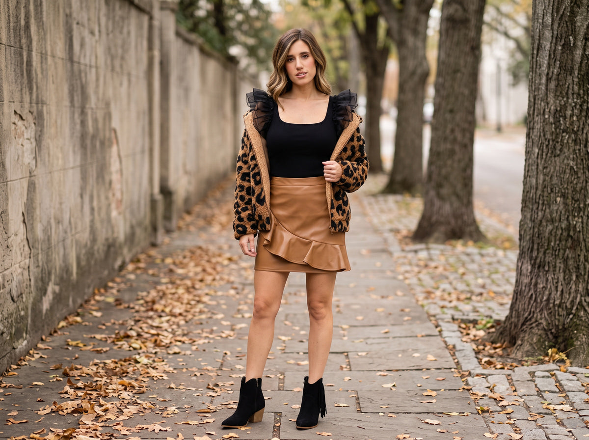 A woman in a black ruffle bodysuit, camel faux leather ruffle mini skirt, leopard sherpa jacket and black fringe boots