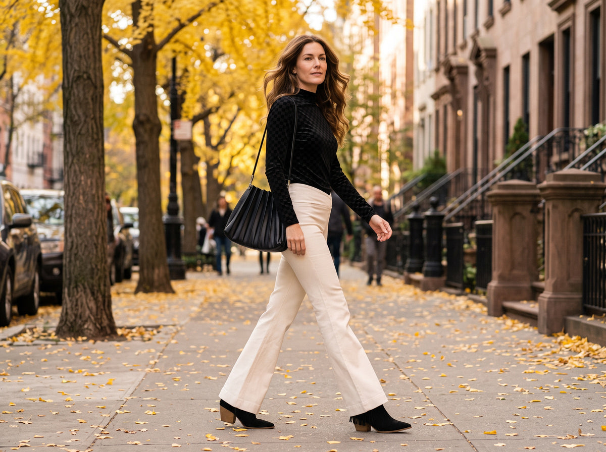 A woman in a black velvet checker mock-neck top, cream flare jeans, black tassel ankle boots and a black pleated shoulder bag walks under golden ginkgo trees