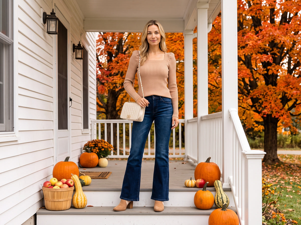 A woman in a camel square-neck sweater, dark flare jeans, tan booties and a cream quilted bag stands on a farmhouse porch with pumpkins and orange maples