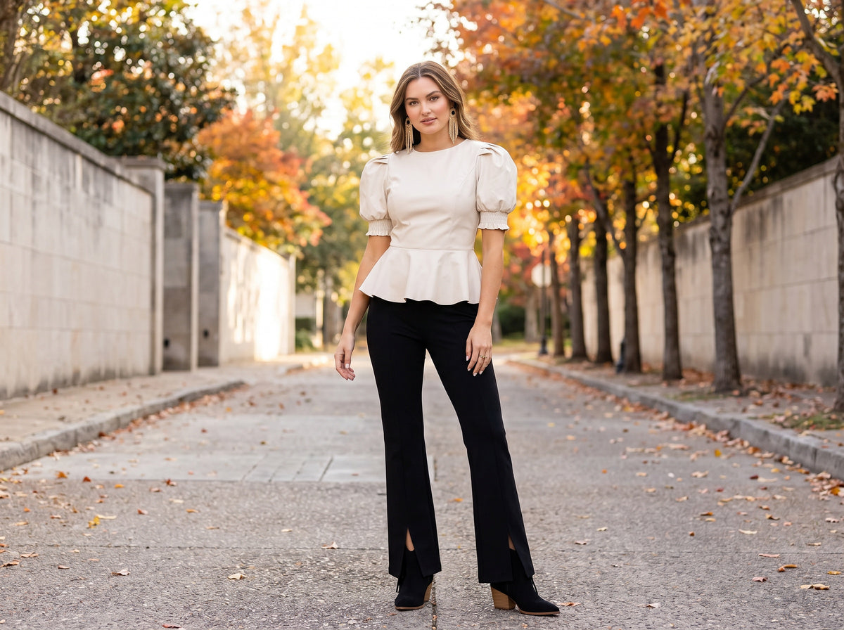 A woman in a cream faux leather puff-sleeve peplum top, black slit flare pants, black fringe boots and gold fringe earrings