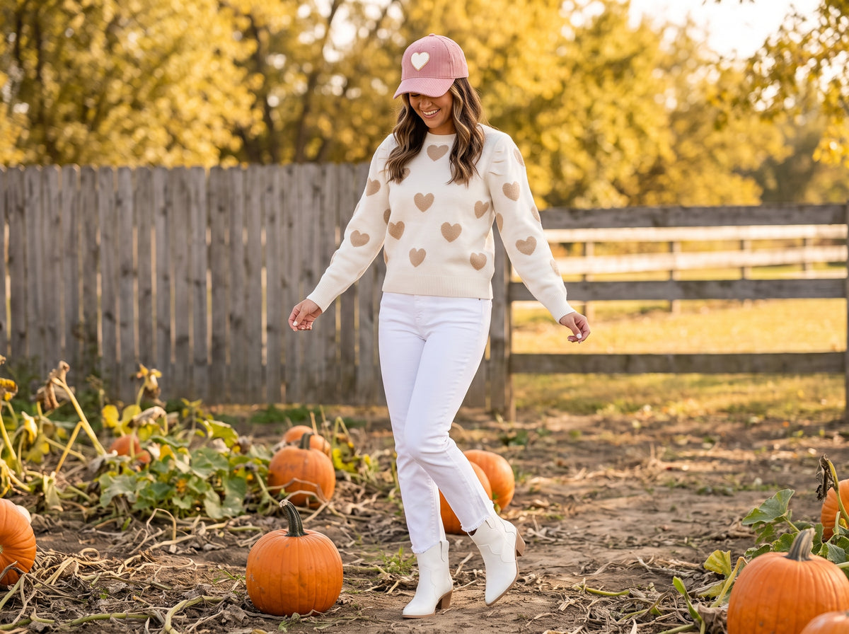 A woman in a cream sweater with gold hearts, white cropped jeans, white cowboy boots and a pink cap in a pumpkin patch