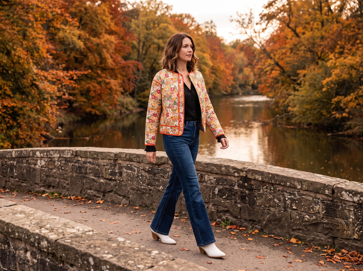 A woman in a floral quilted jacket over a black top, dark flare jeans and white booties crosses a stone bridge over a river lined with autumn trees