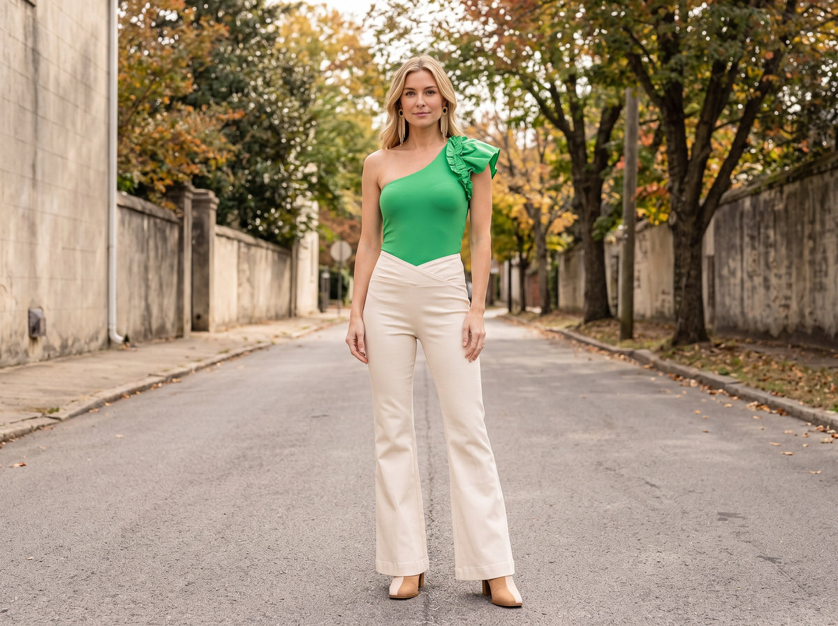 A woman in a green ruffle one-shoulder bodysuit, cream flare jeans, tan booties and gold fringe earrings
