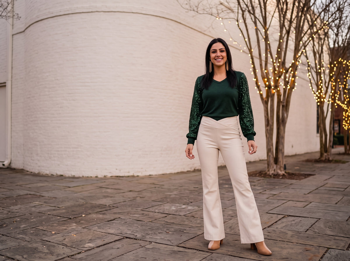 A woman in a green sweater with sequin sleeves, cream flared jeans and suede ankle boots by a white wall