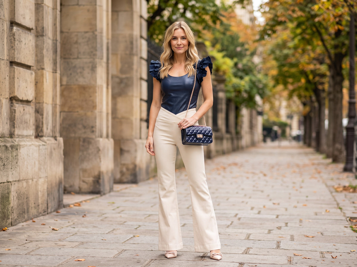 A woman in a navy vegan leather ruffle-sleeve bodysuit, cream flare jeans and nude pumps with a navy quilted bag