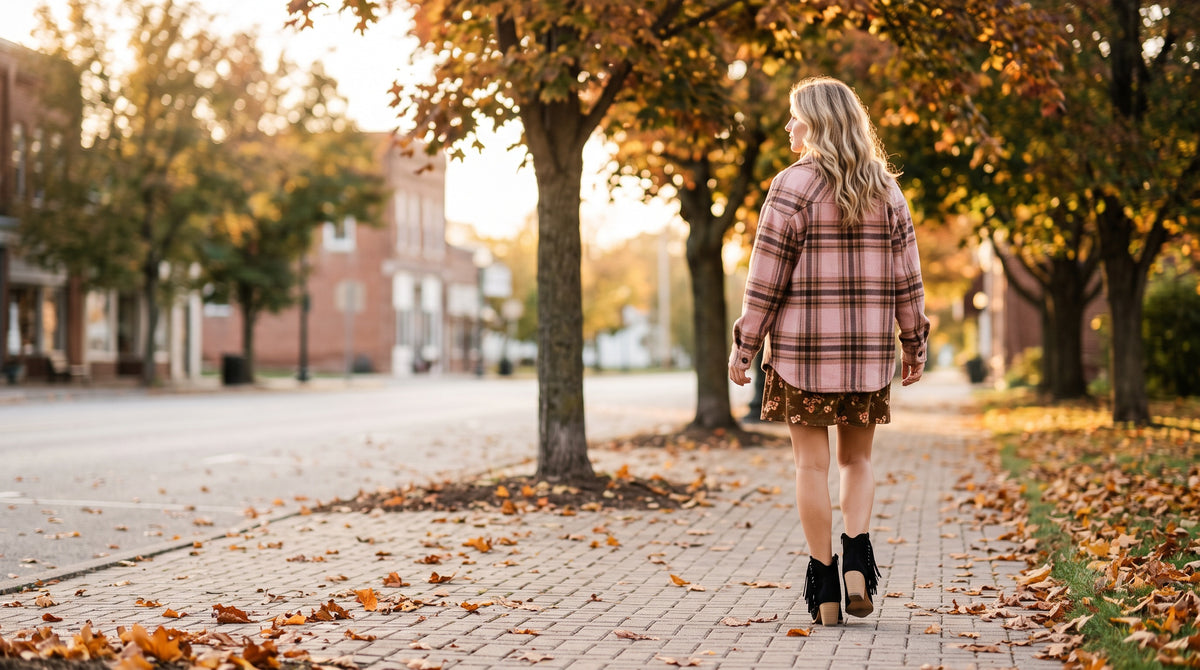 A woman in a pink plaid shacket, floral corduroy mini dress and black tassel ankle boots walks down a leafy autumn sidewalk in golden light