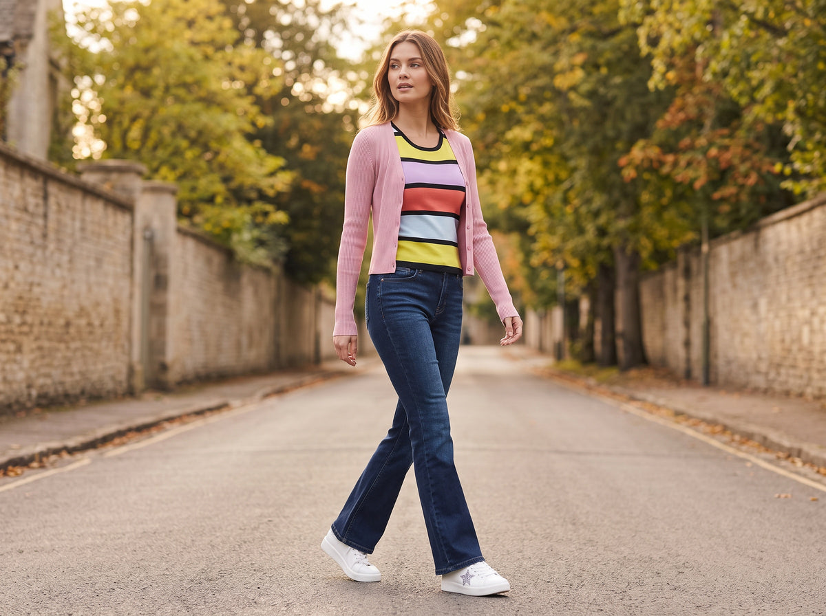 A woman in a pink ribbed cardigan over a striped tank, flare jeans and white star sneakers on an autumn street