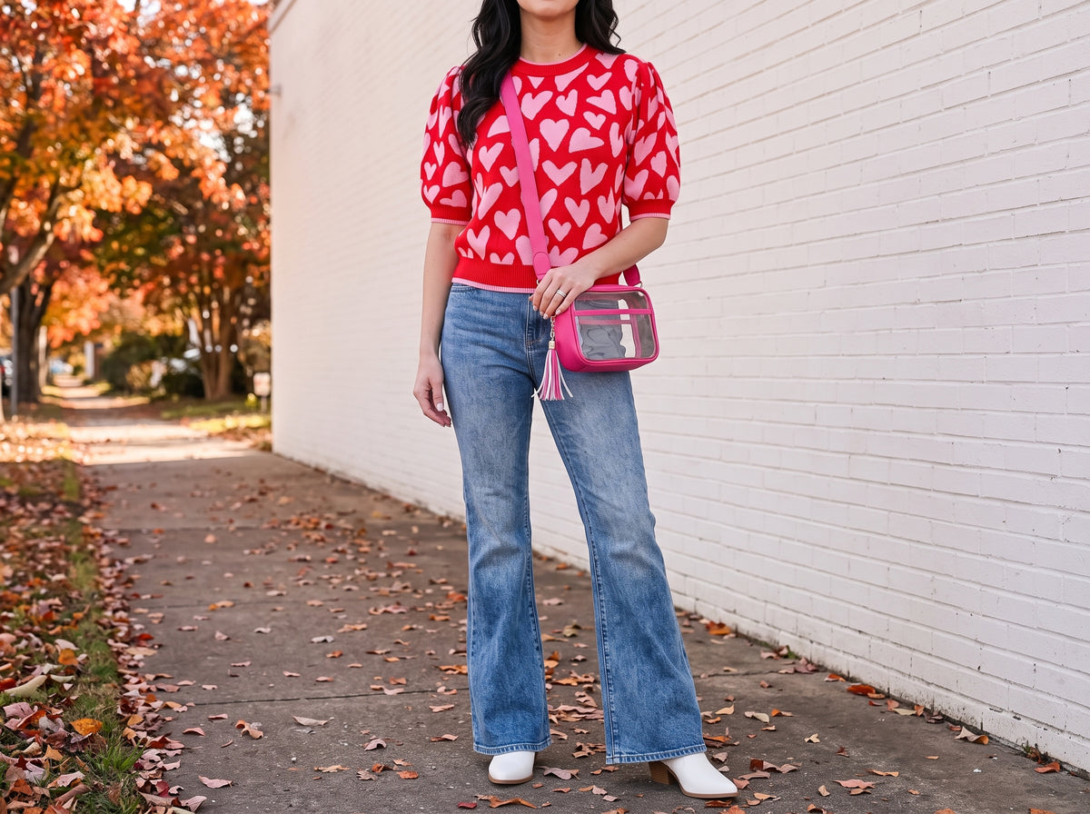 A woman in a red and pink heart puff-sleeve sweater, flare jeans, white cowboy boots and a clear pink crossbody bag