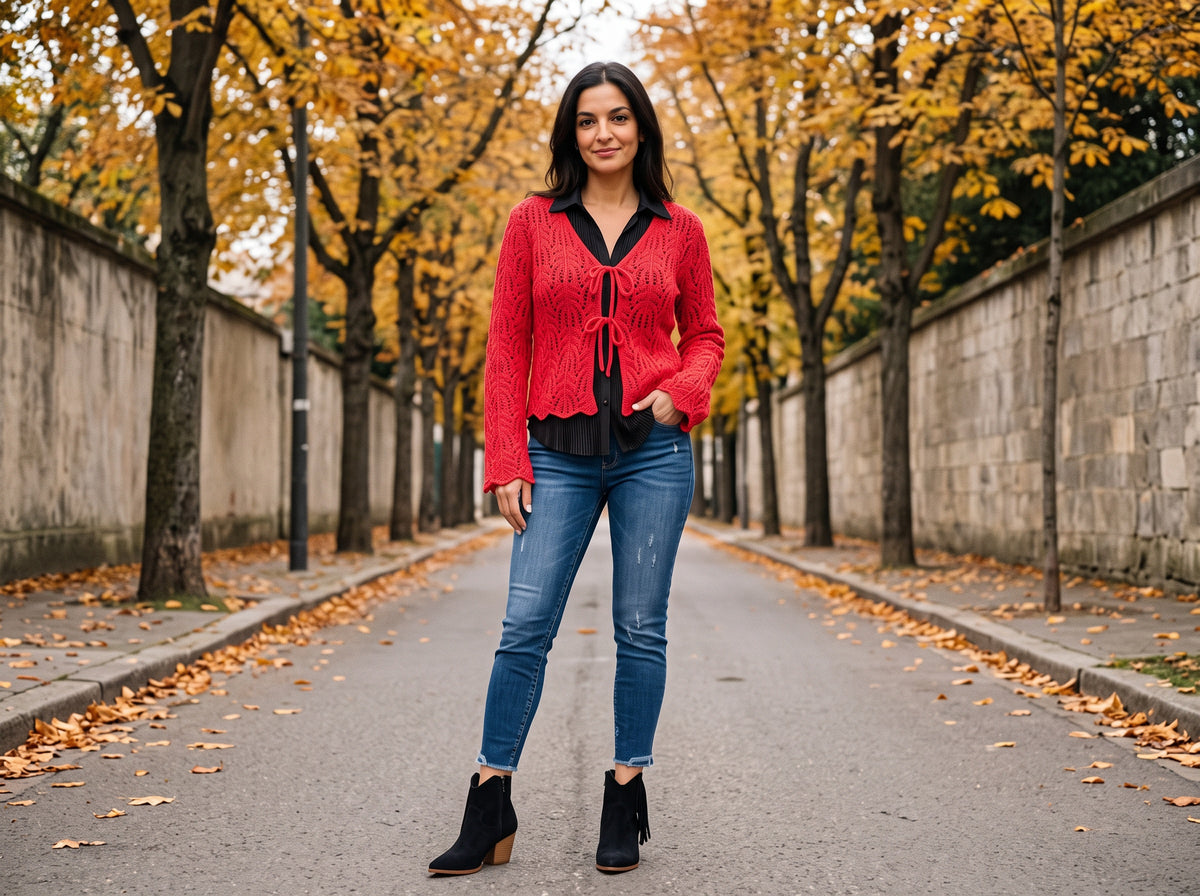 A woman in a red crochet tie-front cardigan over a black top, blue ankle jeans and black fringe ankle boots on an autumn street
