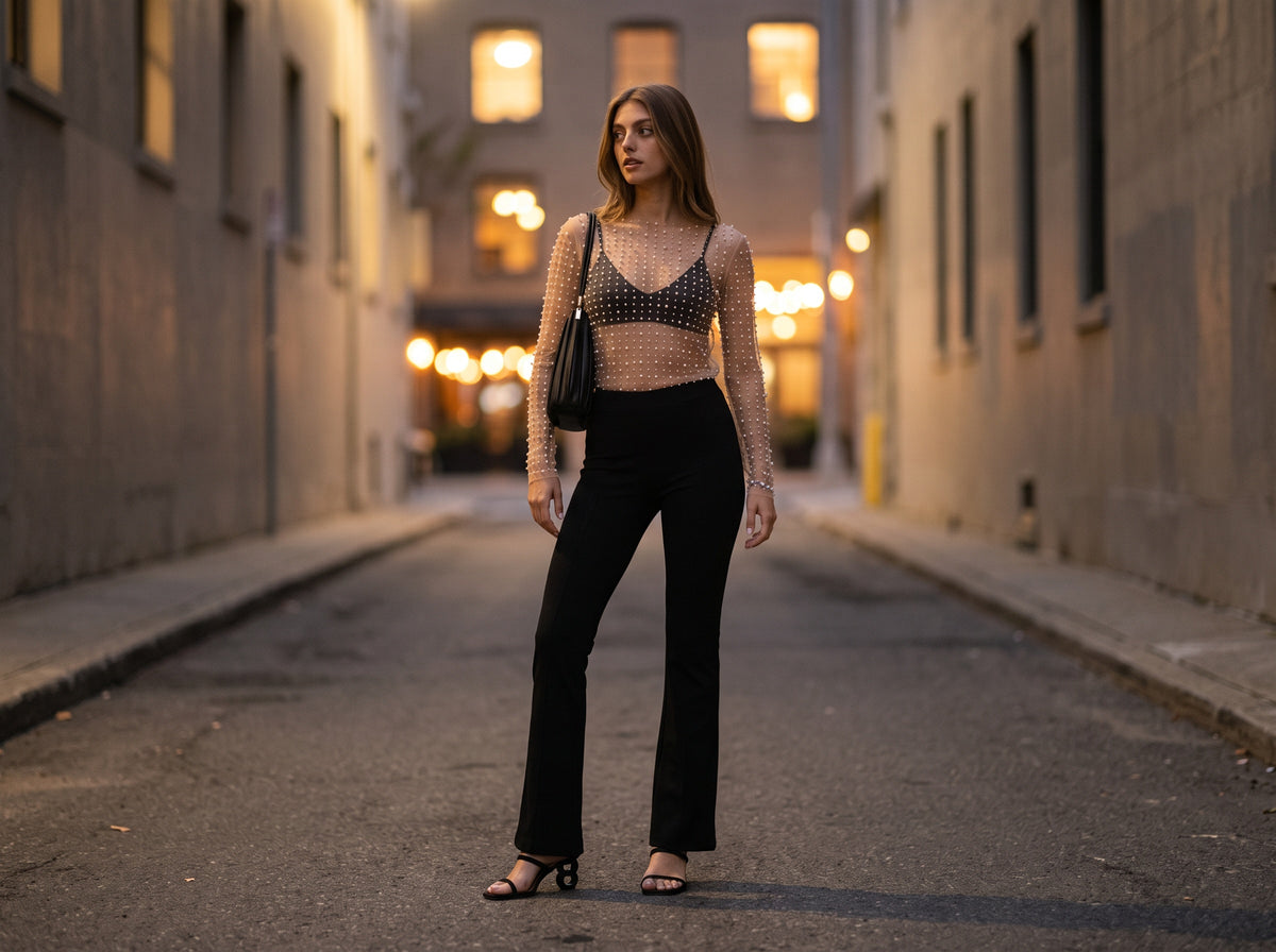 A woman in a sheer pearl-studded mesh top over a black bralette, black flare pants and black heeled sandals at dusk