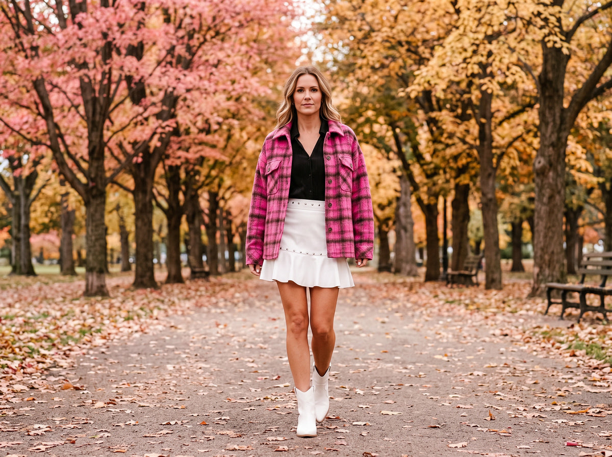 A woman in a white studded mini skirt, a black top, a pink plaid jacket and white western booties walks down an avenue of pink and gold autumn trees