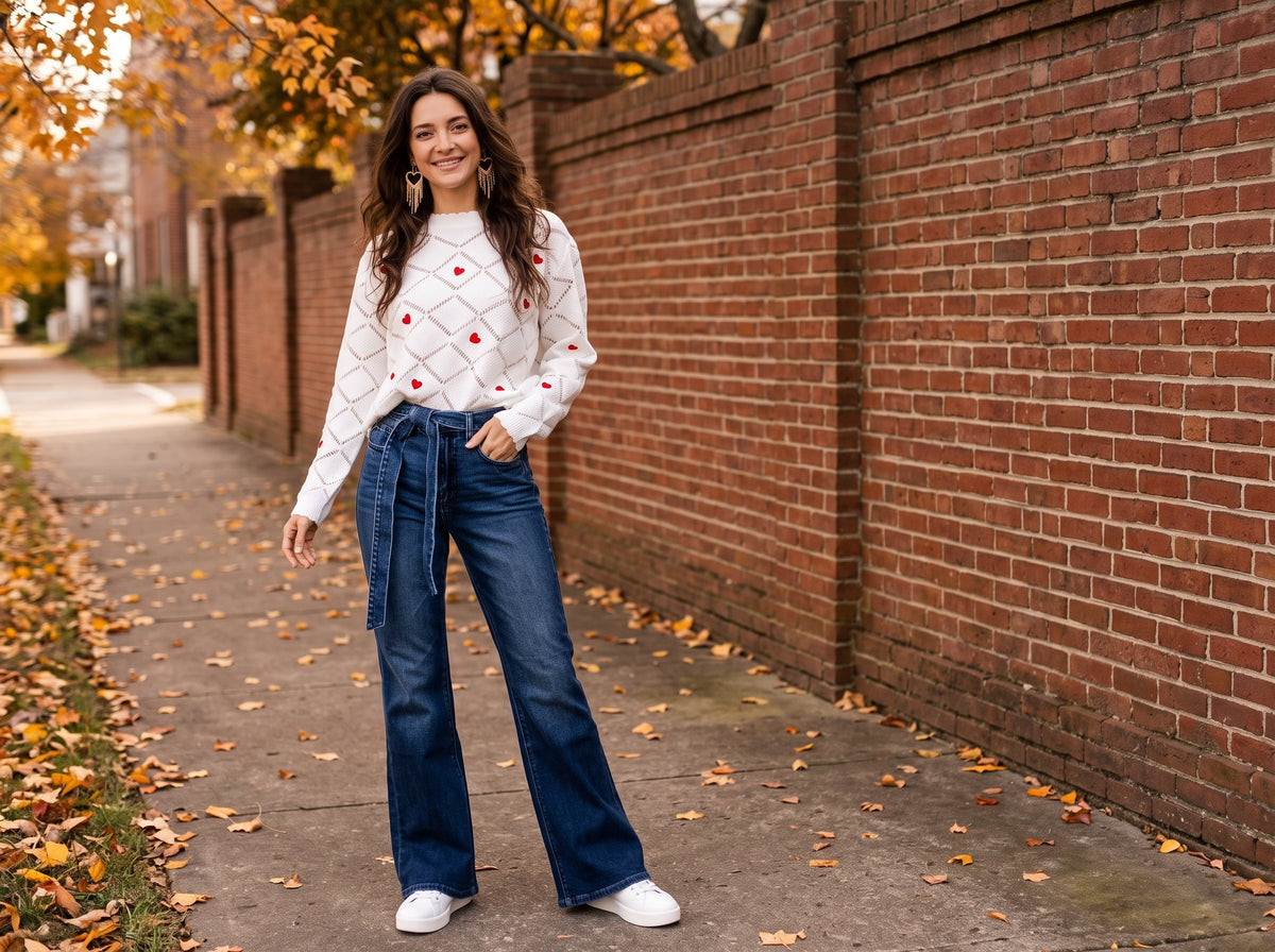 A woman in a white sweater with red hearts, flare jeans and white sneakers beside a brick wall