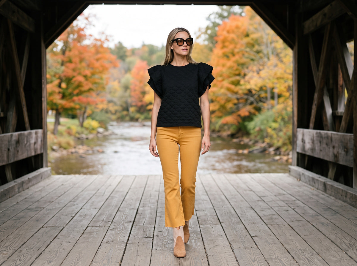 A woman in mustard kick flare pants, a black ruffle-sleeve top, tan booties and oversize sunglasses walks across a covered wooden bridge with autumn trees behind