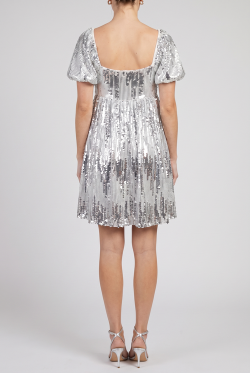 Back view of the silver sequin babydoll mini dress showing the low square back and gathered skirt, on a light-grey studio backdrop.