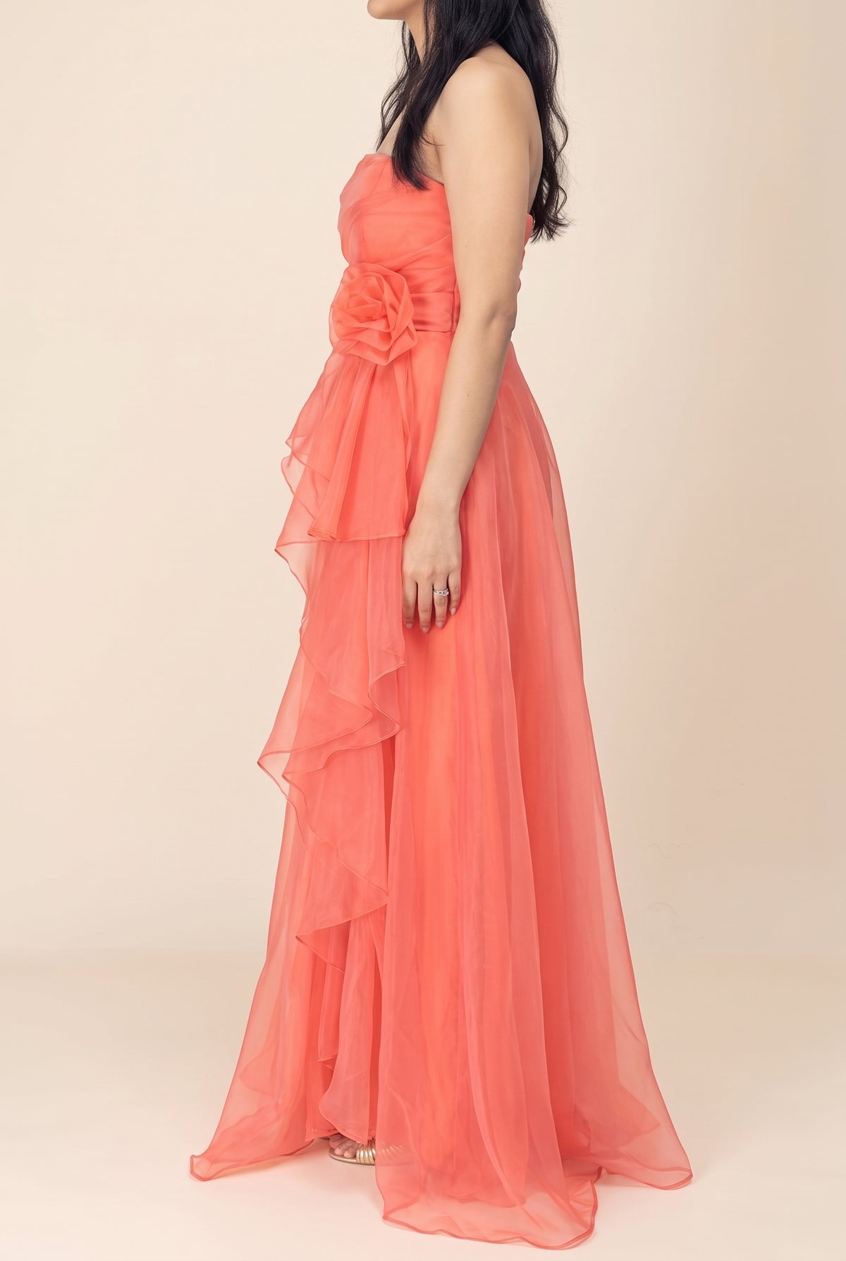Side view of the coral organza maxi dress showing the waist rosette and the cascading front ruffle