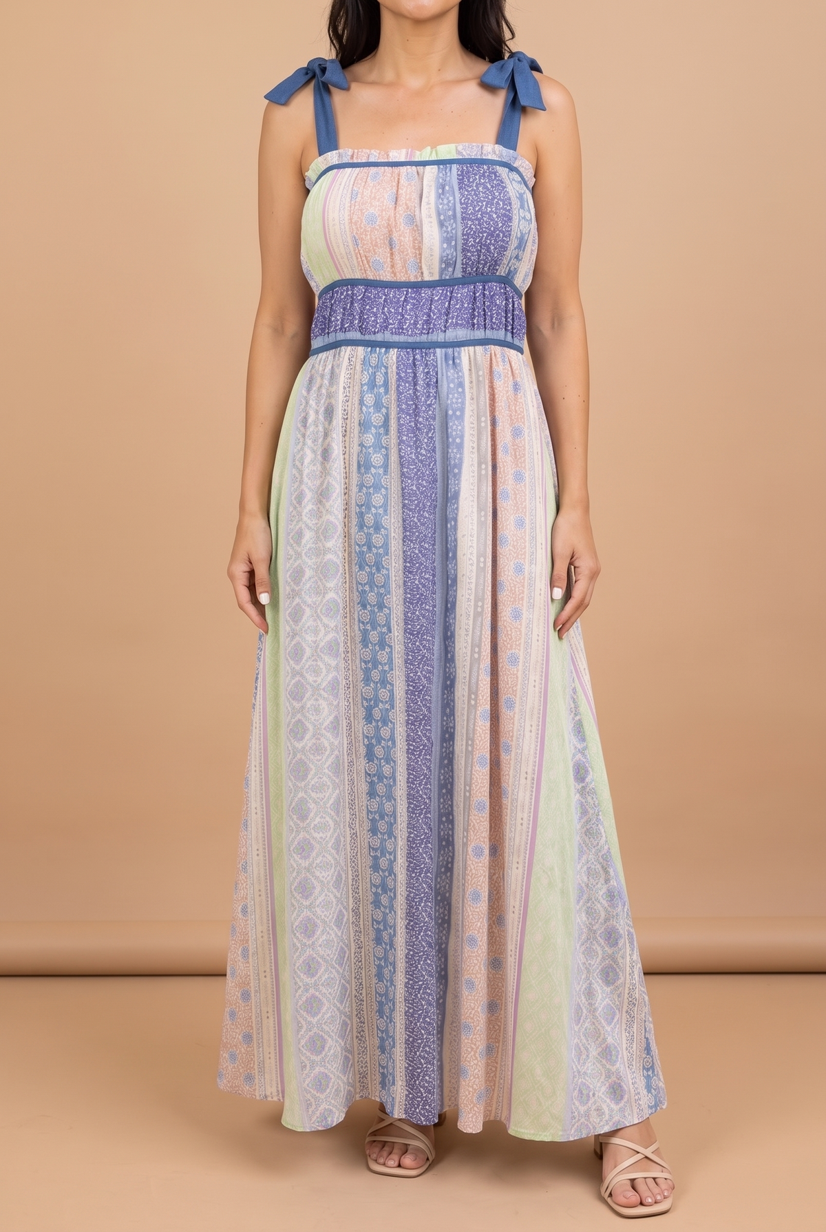 Front view of the Feeling The Sunshine maxi dress on a beige studio backdrop: patchwork print panels, denim-blue tie straps, shirred waist, ankle-length hem.