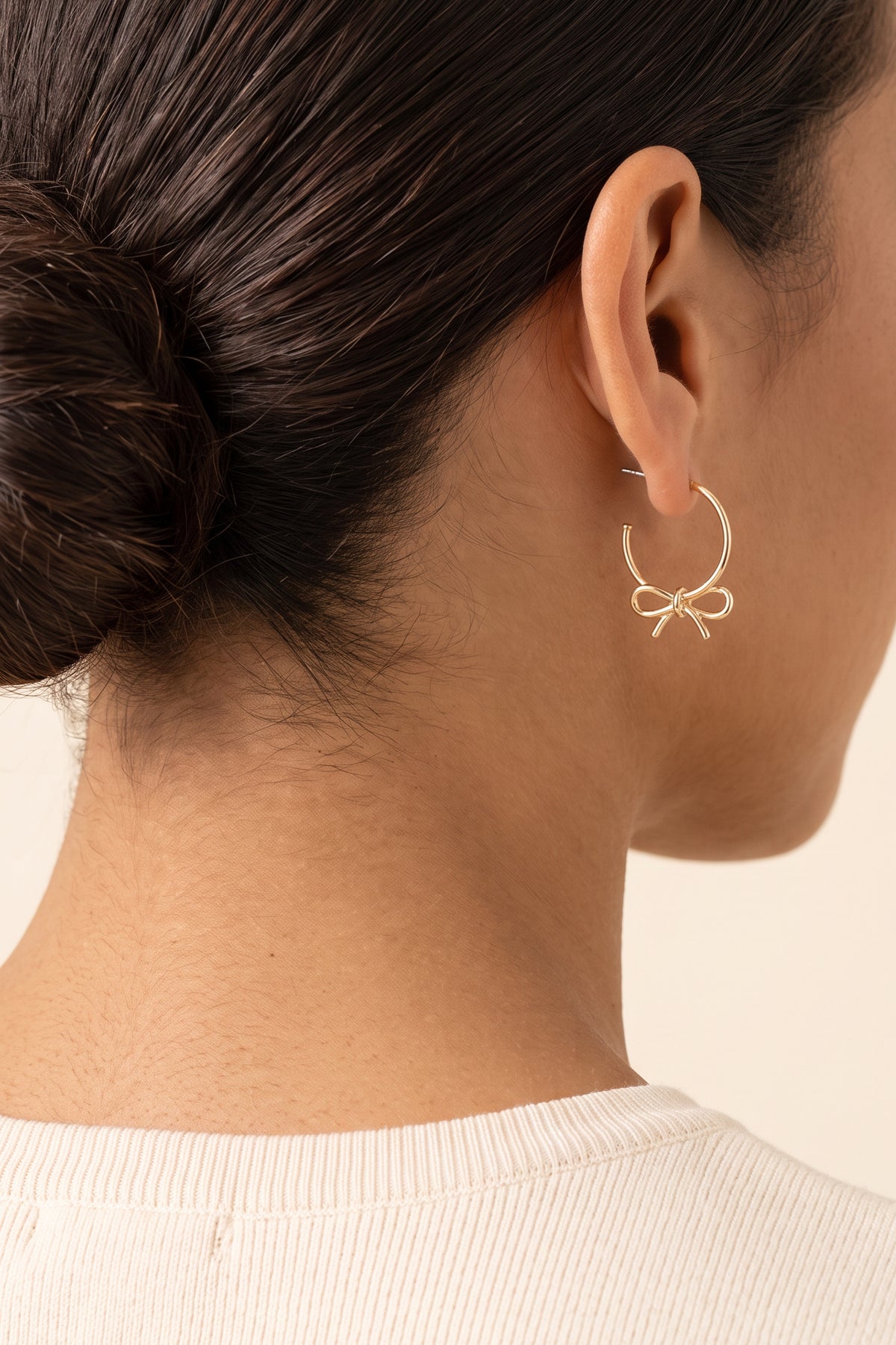Harley Bow Tie Hoop Earrings worn on the ear at true size: a small gold hoop about 1 in across with the bow just below the earlobe