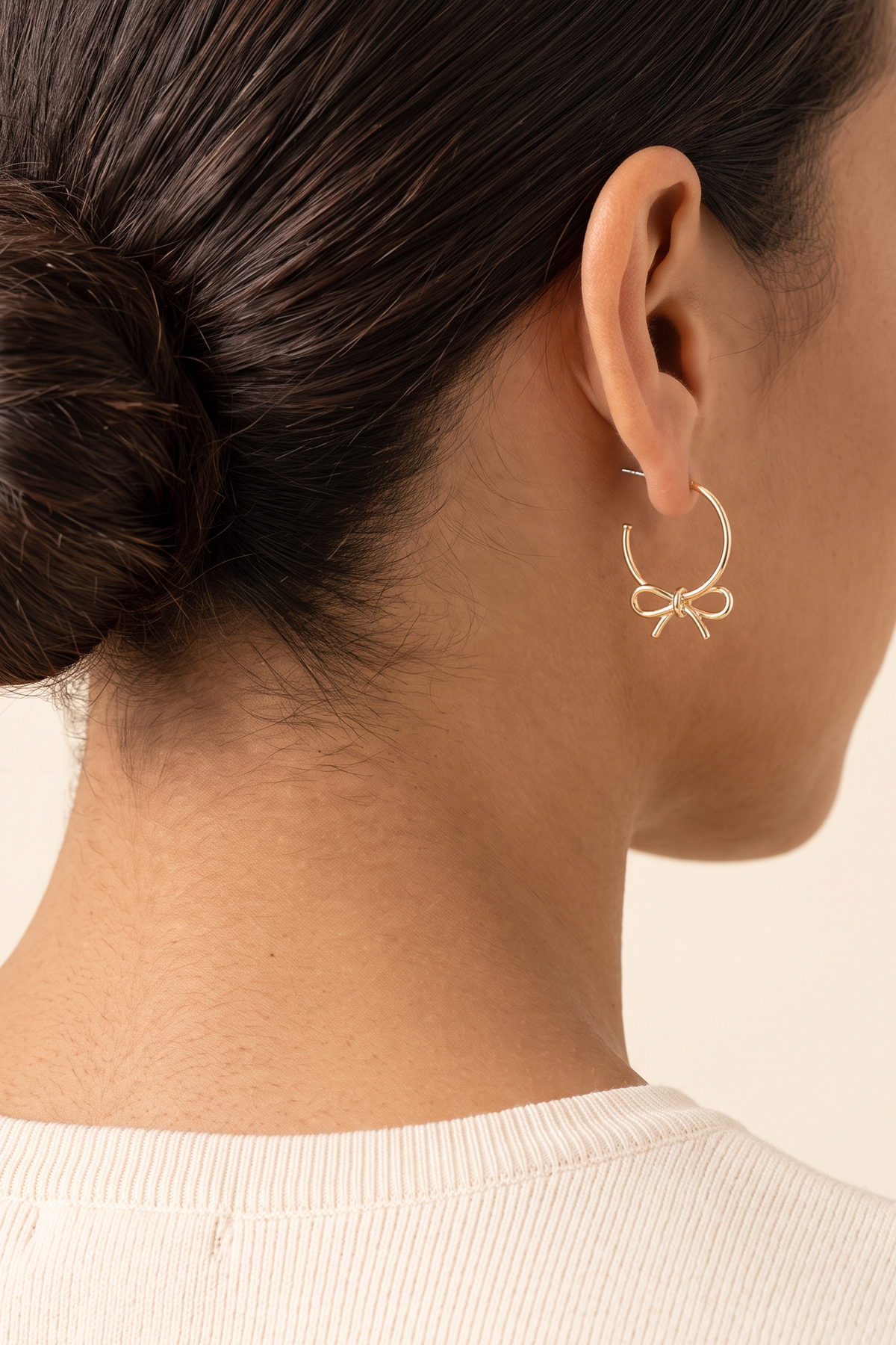 Harley Bow Tie Hoop Earrings worn on the ear at true size: a small gold hoop about 1 in across with the bow just below the earlobe