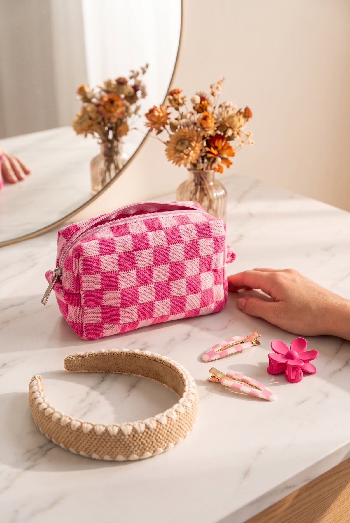 Pink check knit cosmetic pouch on a marble vanity with pink gingham snap clips, a pink flower claw clip and a woven straw headband, a hand beside it for scale
