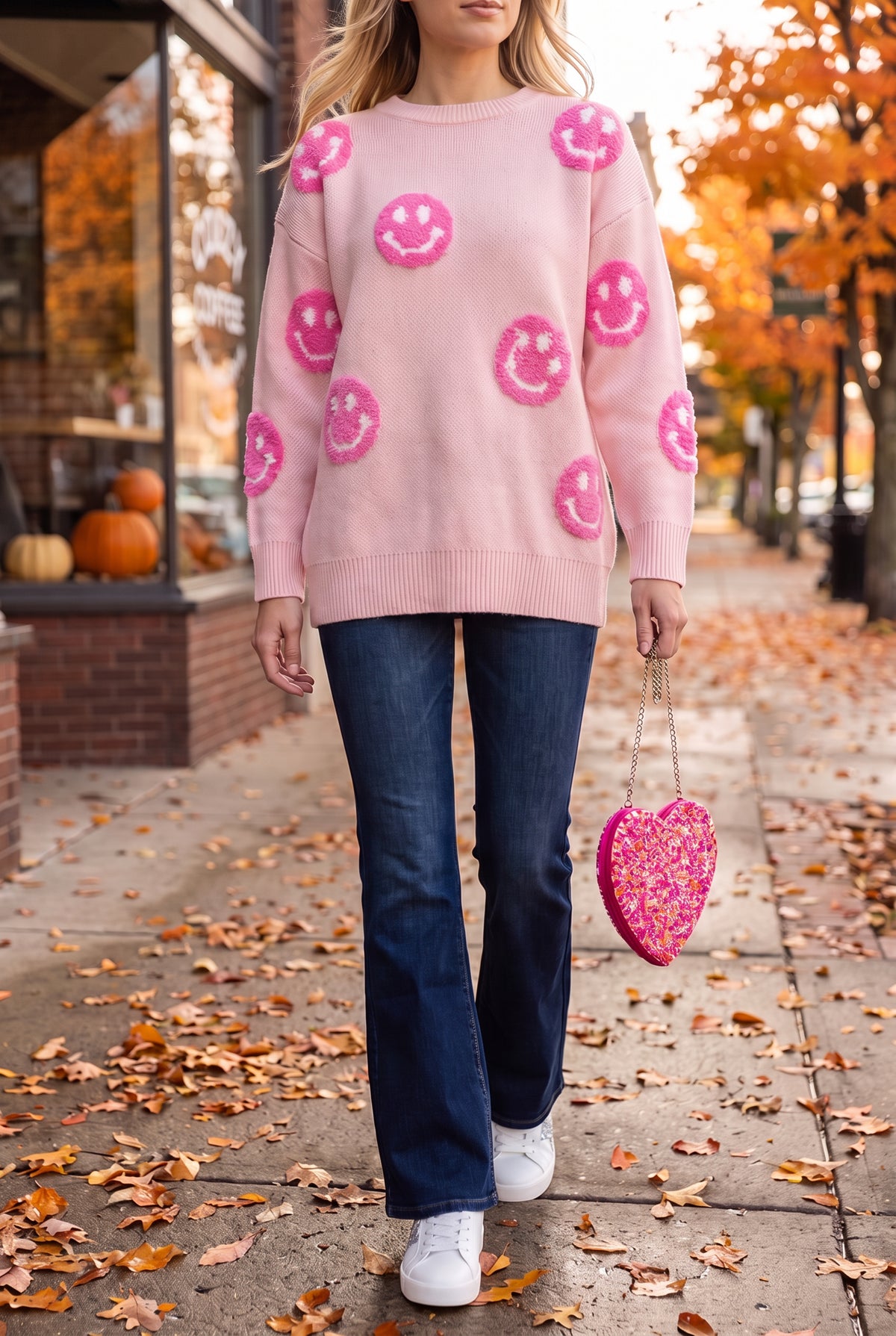 Pink happy face sweater worn with dark flare jeans, white glitter star sneakers and a pink beaded heart bag on an autumn sidewalk.