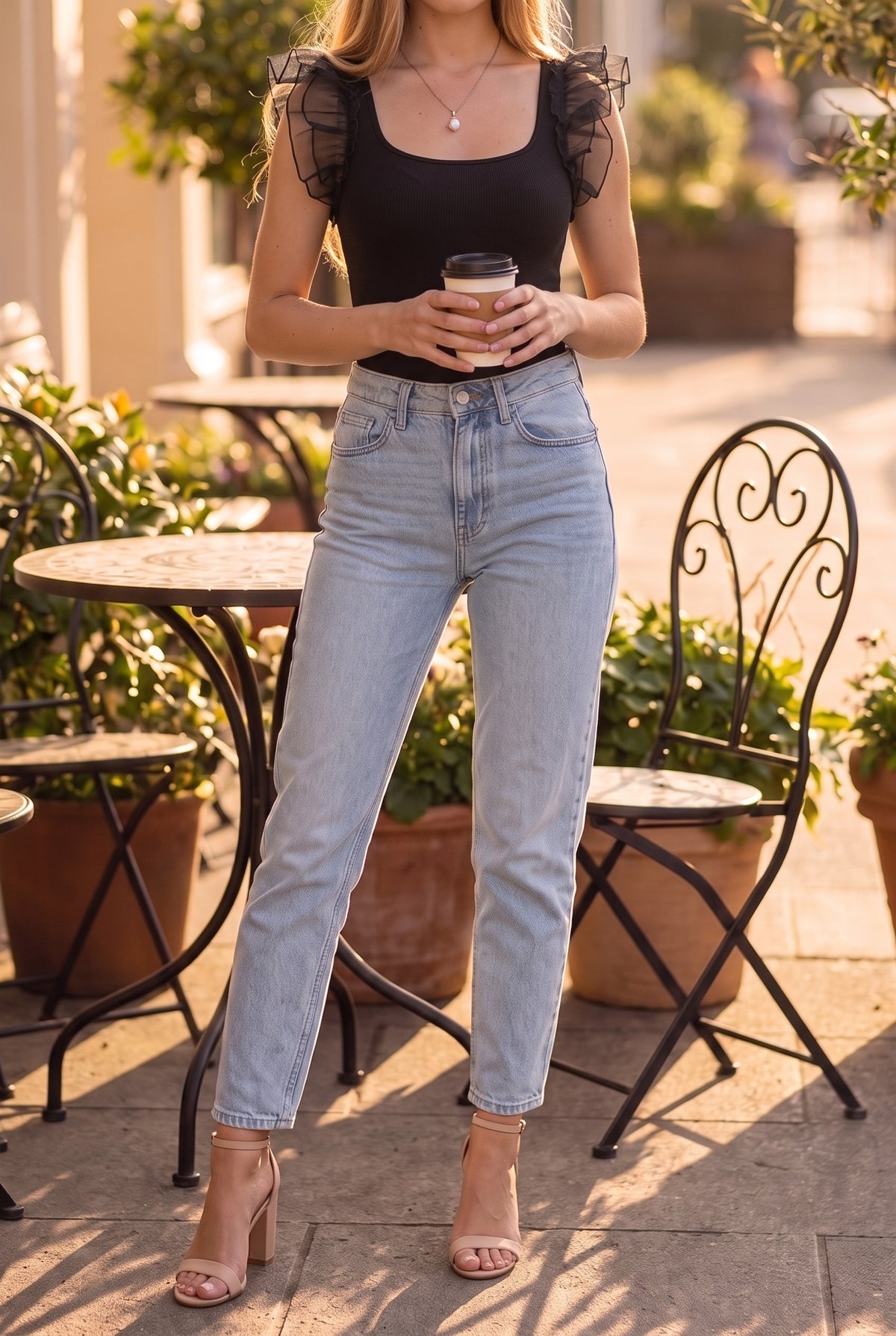 Model in the black square-neck ruffle sleeve bodysuit and light jeans holding a coffee on a café terrace, from the chin down