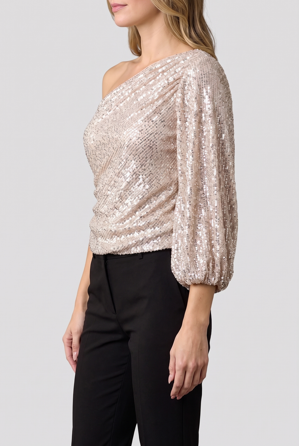 One-shoulder sequin top worn with black trousers, side view on a light-grey studio backdrop