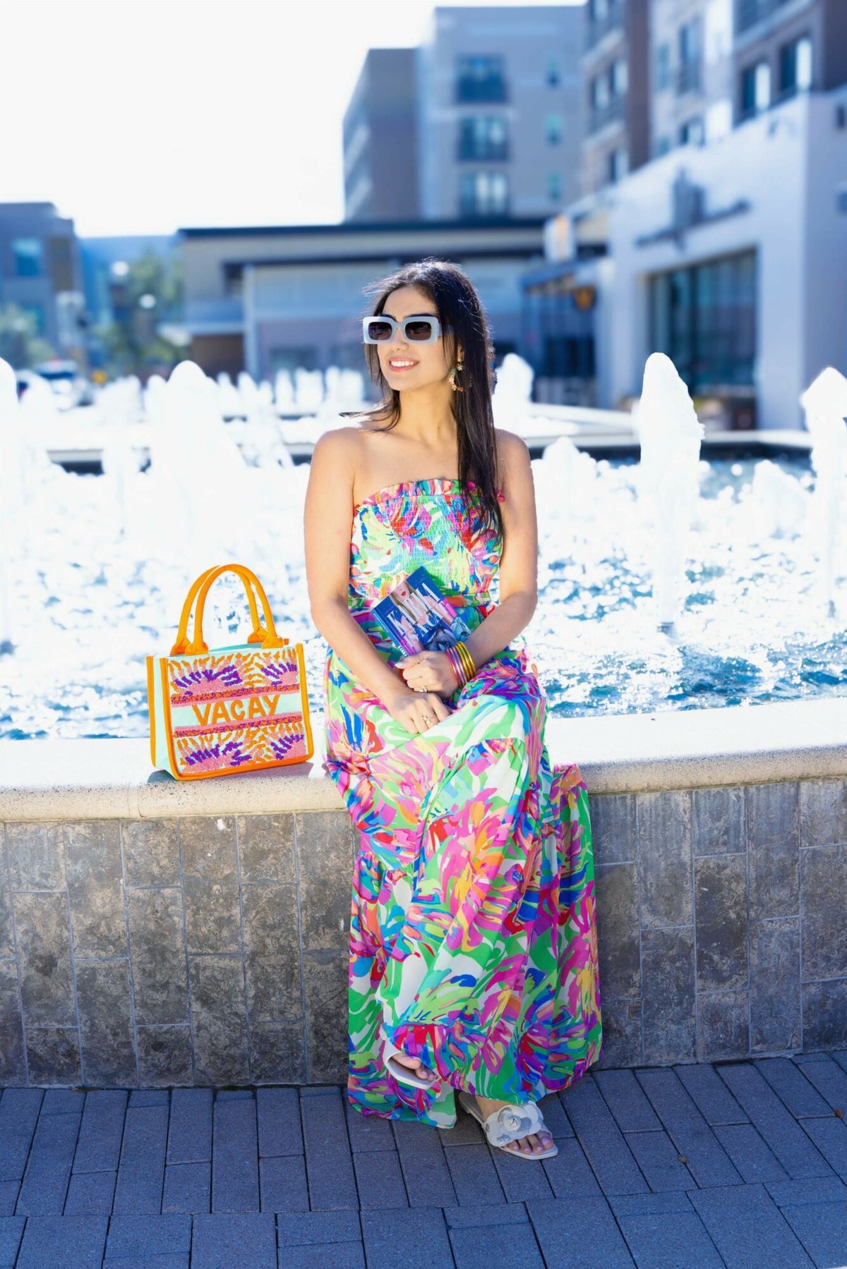 Shweta in a bright tropical maxi dress sitting by a fountain with a beaded Vacay tote