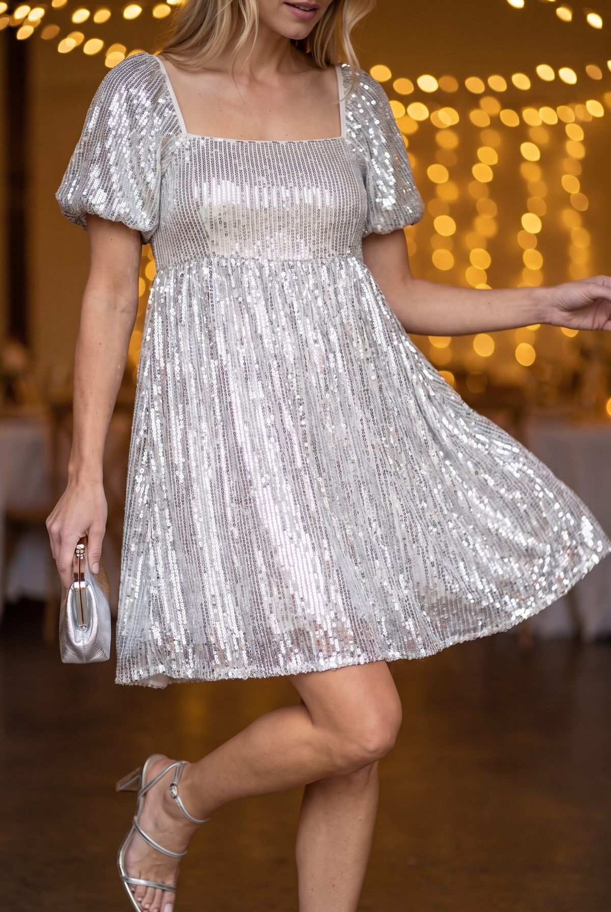 The silver sequin babydoll mini dress worn at an evening party with string lights behind, a small silver clutch and silver strappy heels.