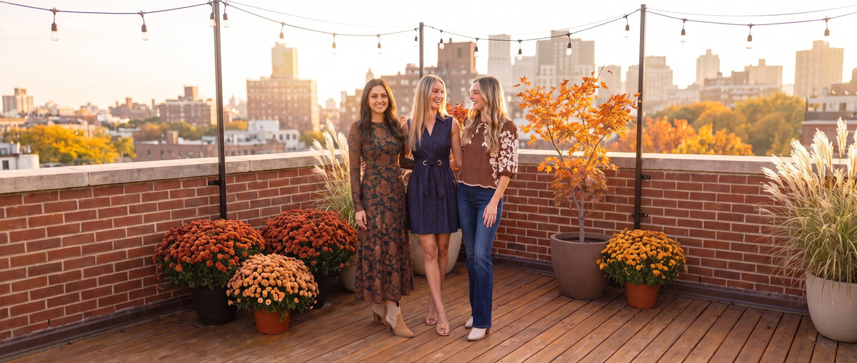 Three women laughing together on a wide sunny autumn city rooftop with string lights and potted mums, one in a green and rust lace maxi dress, one in a navy belted mini dress, one in an embroidered brown top and jeans