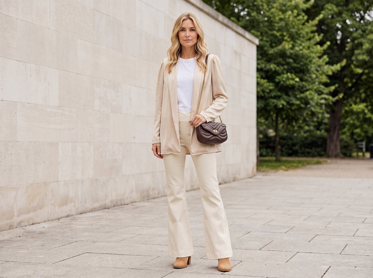 Woman in a beige blazer, white top and cream flared jeans with a brown quilted chain crossbody bag and suede ankle booties
