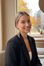 Woman in a black blazer at a cafe, hair tucked behind her ear to show the gold fringe earrings