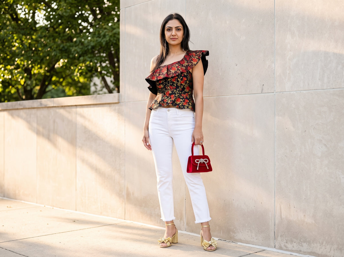 Woman in a black floral one-shoulder crop top with white cropped jeans, gold bow sandals and a red velvet bag