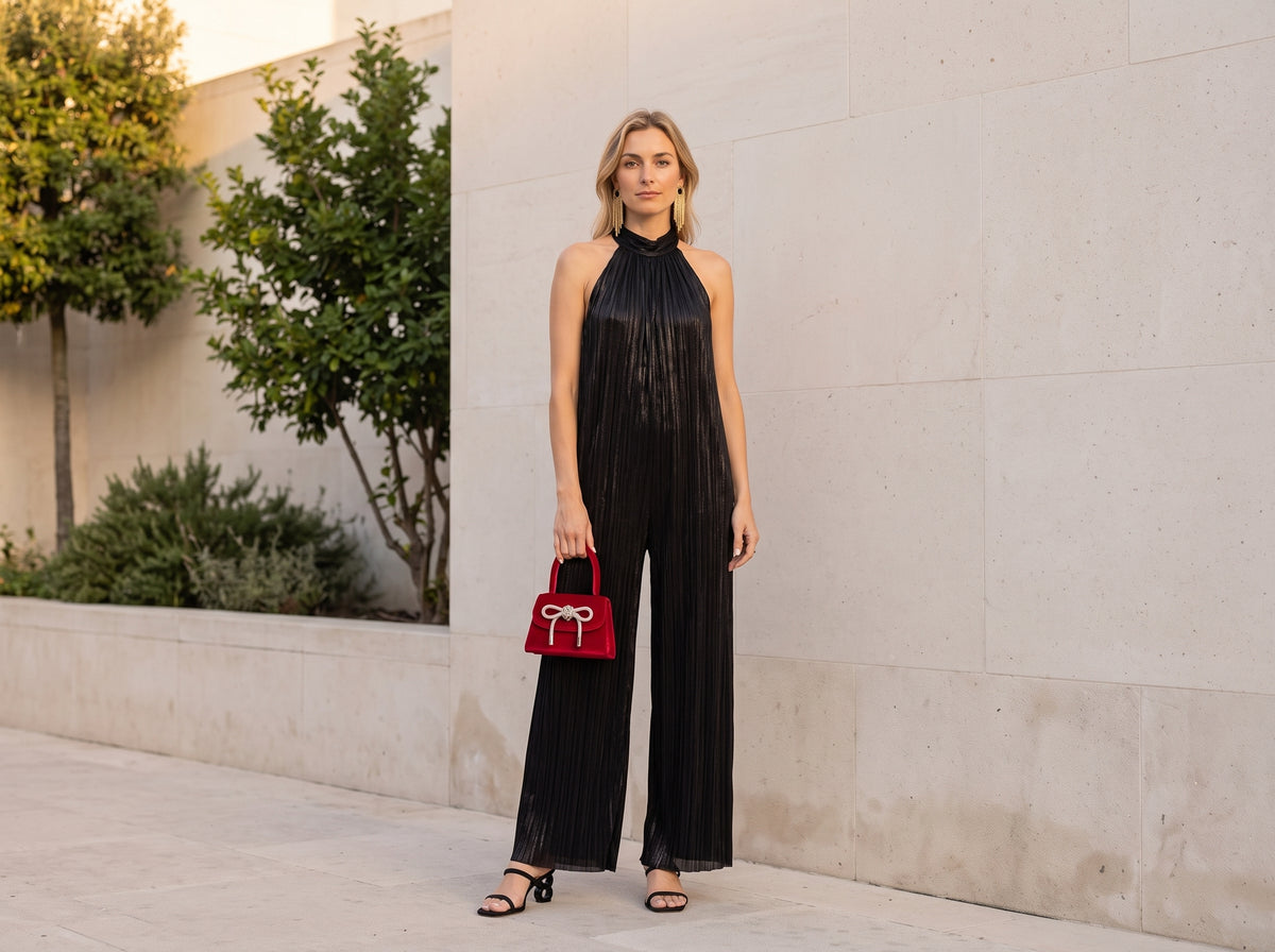 Woman in a black halter-neck wide-leg jumpsuit and heeled sandals holding a red velvet mini bag with a bow