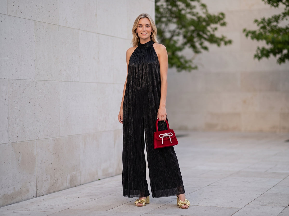 Woman in a black halter-neck wide-leg jumpsuit with gold bow sandals, a red velvet bow bag and gold hoops