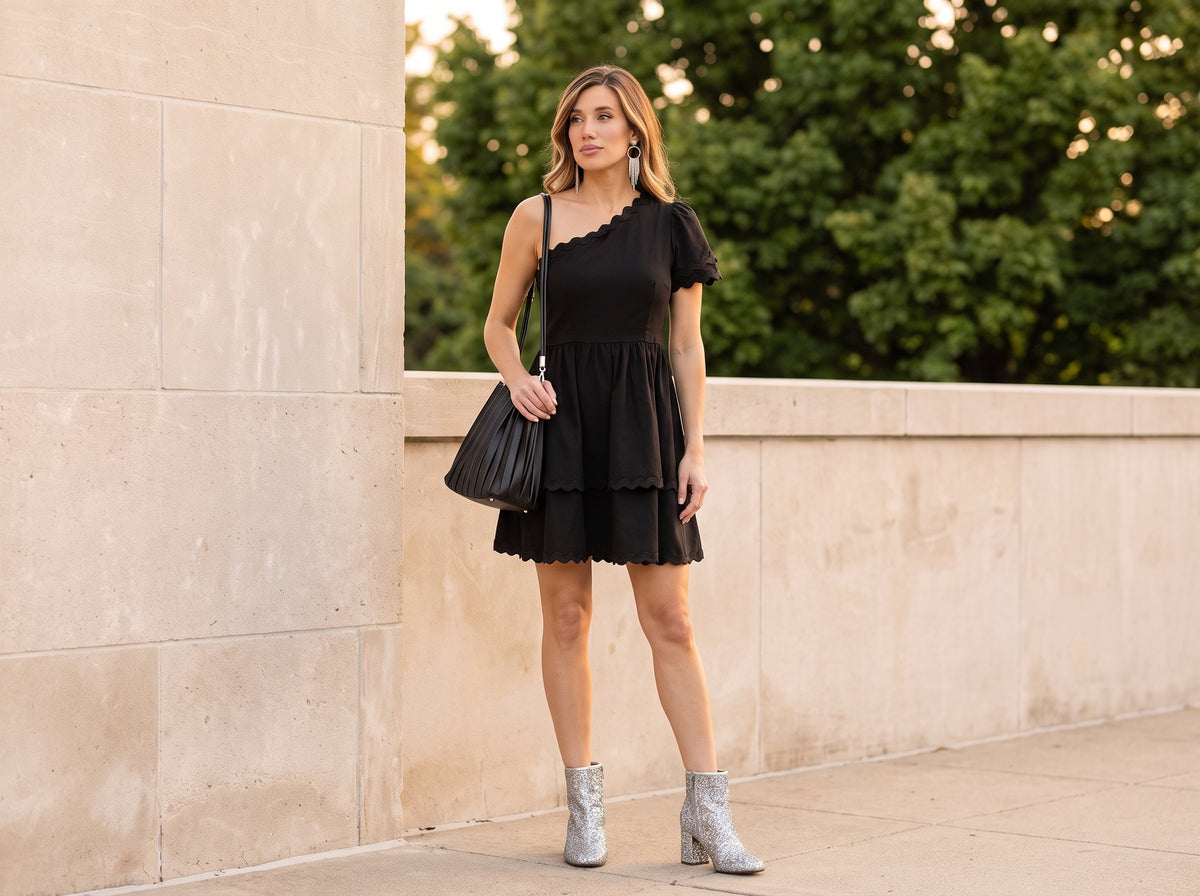 Woman in a black one-shoulder dress with a black pleated shoulder bag, silver chandelier earrings and silver glitter ankle boots