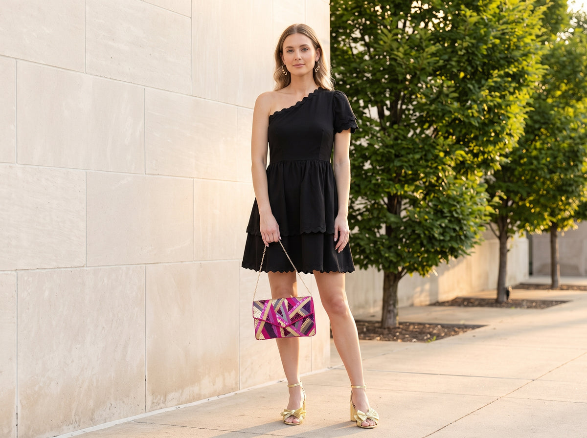 Woman in a black one-shoulder mini dress and gold bow heeled sandals holding a pink beaded party clutch