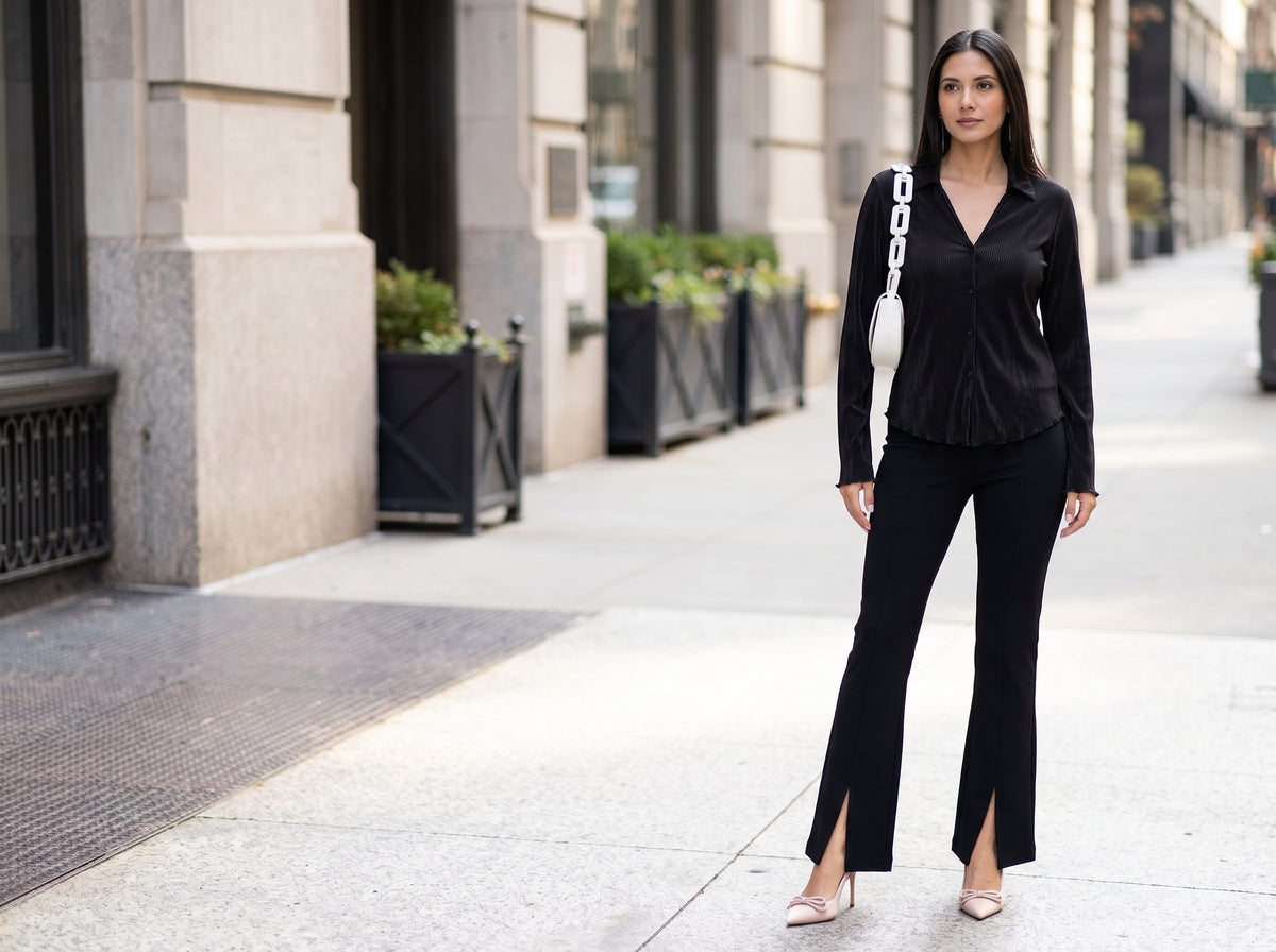 Woman in a black pleated shirt and black flared pants on a city street with a white shoulder bag on a chain strap