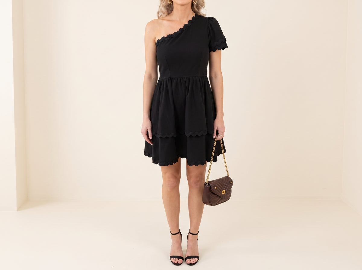 Woman in a black scalloped one-shoulder tiered mini dress with a quilted chain bag