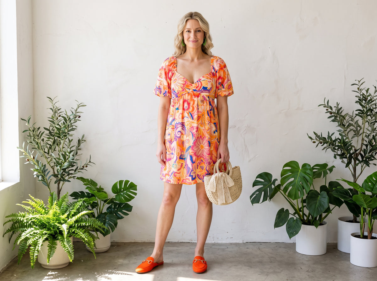Woman in a bright floral A-line mini dress with orange suede mules, a straw crossbody bag and woven hoop earrings