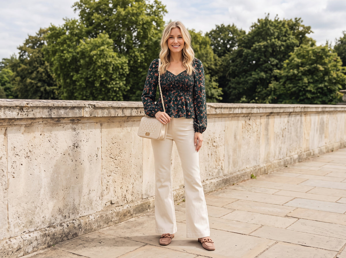 Woman in a floral long-sleeve blouse and cream flared jeans with a beige quilted bag and suede chain mules