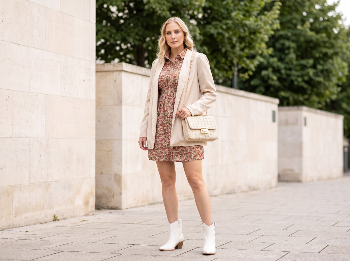 Woman in a floral mini dress and beige blazer with a cream braided chain-strap bag and white cowboy ankle boots