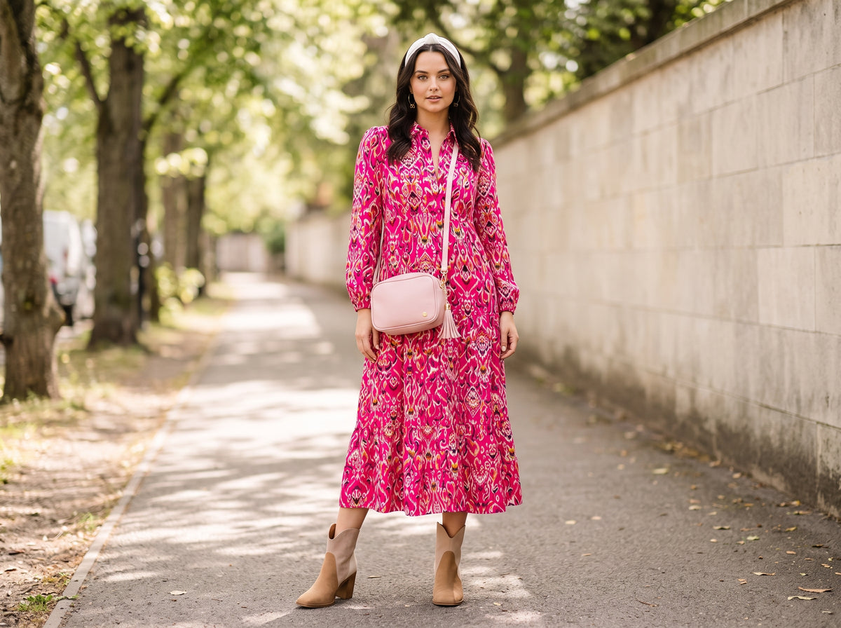 Woman in a fuchsia print midi shirt dress with suede ankle booties, a pink tassel crossbody and gold bow hoop earrings