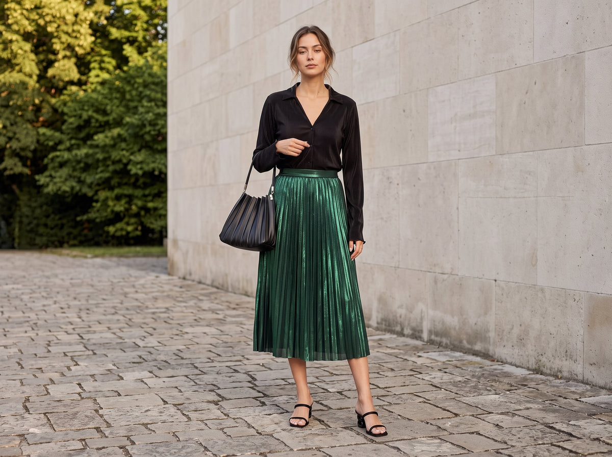 Woman in a green metallic pleated midi skirt with a black pleated top, black heeled sandals and a black bag