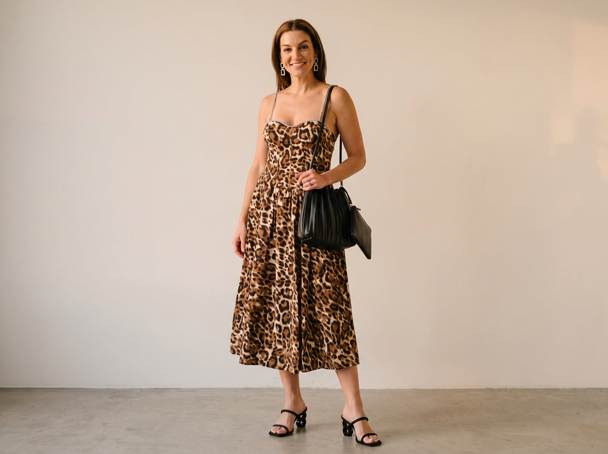 Woman in a leopard print midi dress with rhinestone straps, black heeled sandals, a black pleated shoulder bag and rhinestone earrings
