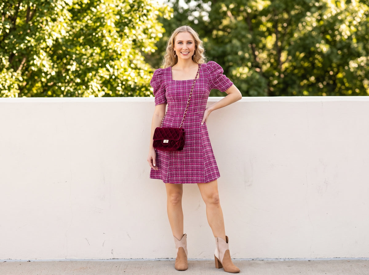 Woman in a magenta check square-neck puff-sleeve dress with suede ankle booties, a burgundy velvet crossbody and gold bow hoops