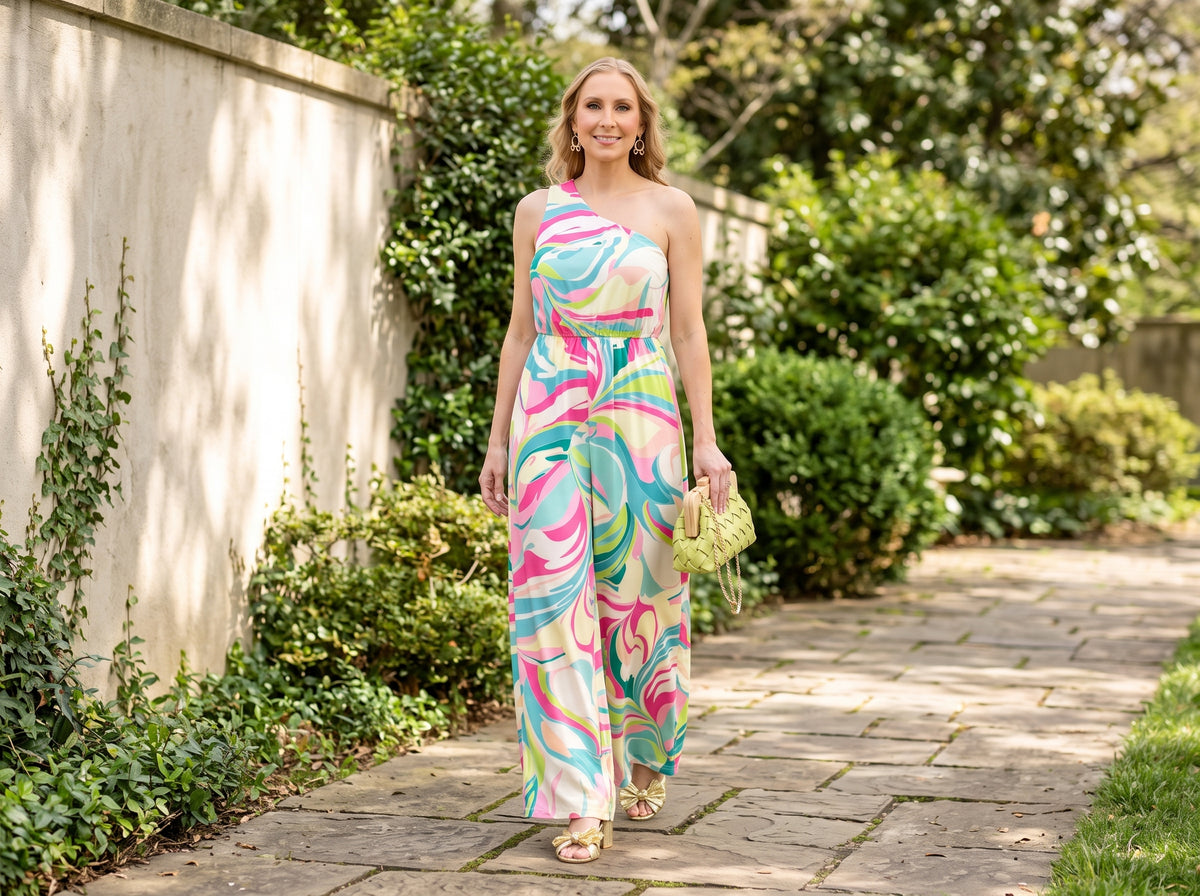 Woman in a one-shoulder swirl-print jumpsuit with gold bow ankle-strap sandals, a woven clutch and gold bow hoop earrings