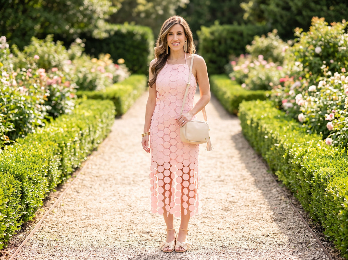 Woman in a pink fringe crochet lace halter midi dress with beige block-heel sandals, a pink tassel bag and gold fringe earrings