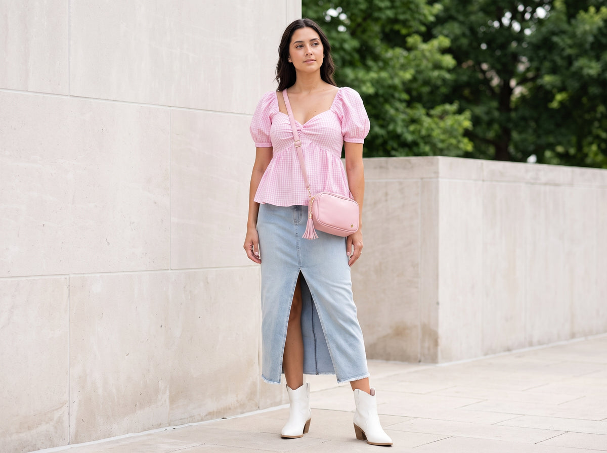 Woman in a pink gingham puff-sleeve top with a denim midi skirt, white western booties and a pink crossbody